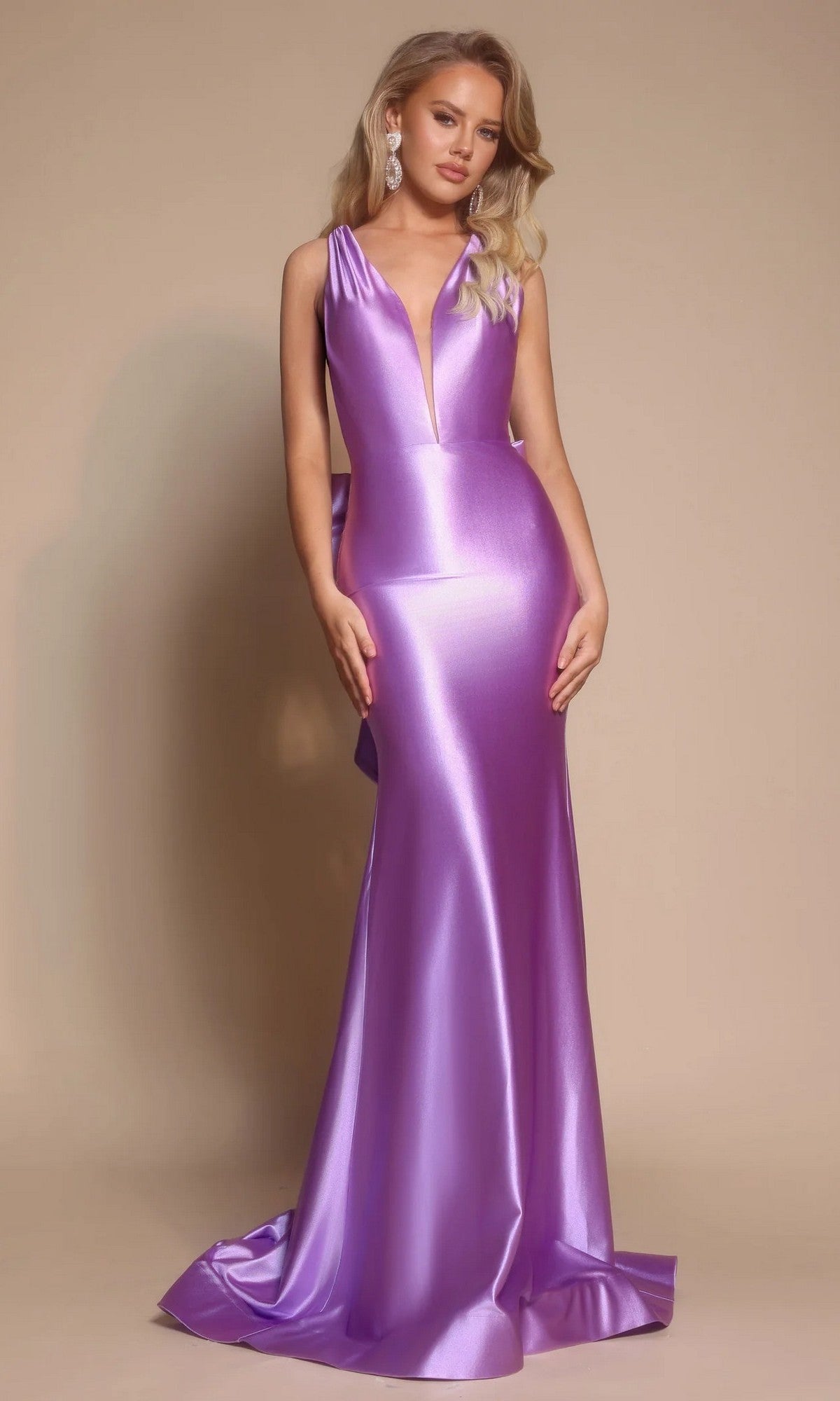 A woman poses against a beige background in the Portia and Scarlett XO095 Long Formal Dress, a sleeveless, floor-length metallic lavender gown with a deep V neckline, open back, and statement earrings.